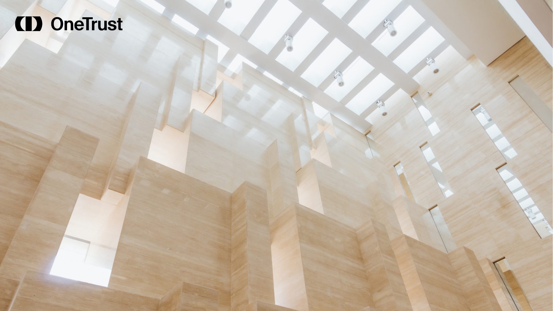 Bright modern atrium with cream stone walls, skylights, and a black OneTrust logo in the upper-left corner.