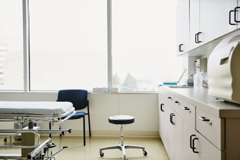 Bright medical examination room with an exam bed, rolling stool, sink, cabinets, and large windows.